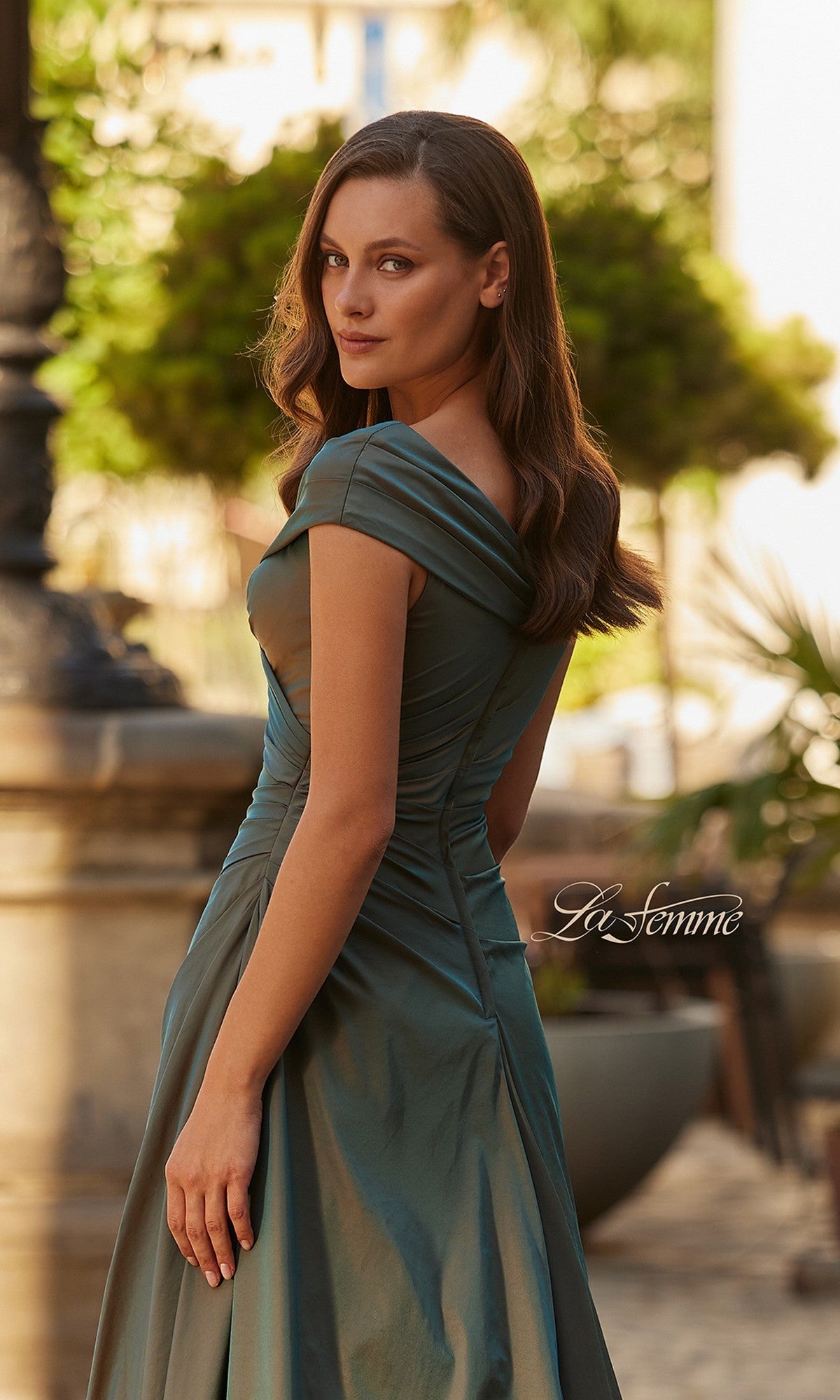 A woman in a green La Femme 33446 long formal dress stands outdoors, glancing over her shoulder. The background shows trees and blurred building elements, with
